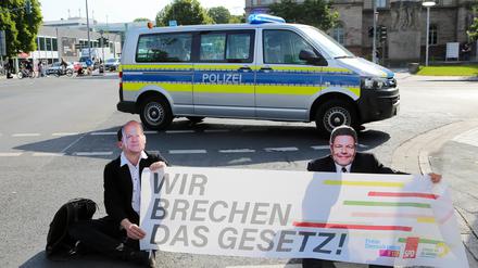Klimaaktivisten der Gruppe Letzte Generation sitzen mit Masken von Bundeskanzler Scholz (l) und Wirtschaftsminister Habeck bei einer Sitzblockade auf einer Kreuzung vor einem Polizeiauto. Die Aktivisten halten ein Banner mit der Aufschrift „Wir brechen das Gesetz!“