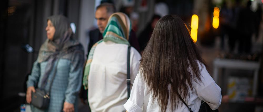 Eine Frau läuft in Teheran mit offenen Haaren am Abend eine Straße entlang, im Hintergrund laufen zwei Frauen mit Kopftuch.