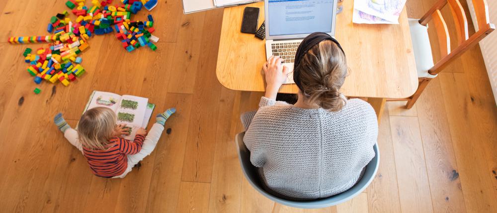 Ein zweijähriges Kind spielt im Wohnzimmer, während seine Mutter im Homeoffice an einem Laptop arbeitet. Immer mehr Mütter minderjähriger Kinder in Deutschland sind erwerbstätig.