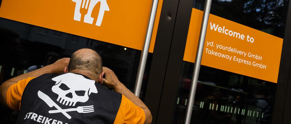 17.08.2023, Berlin: Ein Teilnehmer schaut bei einer Kundgebung zum Streik von Beschäftigten des Essenslieferdienstes Lieferando für bessere Bezahlung durch eine Glastür des Firmensitzes von Lieferando.