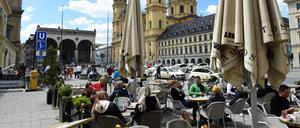 Unter weißblauem Himmel sitzen Menschen im Außenbereich vor dem Cafe-Restaurant Tambosi auf dem Odeonsplatz gegenüber der Theatinerkirche.