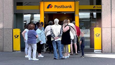 Kunden stehen in einer Warteschlange vor einer Filiale einer Postbank. 