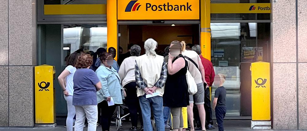 Kunden stehen in einer Warteschlange vor einer Filiale einer Postbank. 