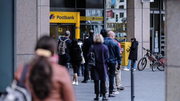 Kunden stehen in einer Warteschlange vor einer Filiale einer Postbank.