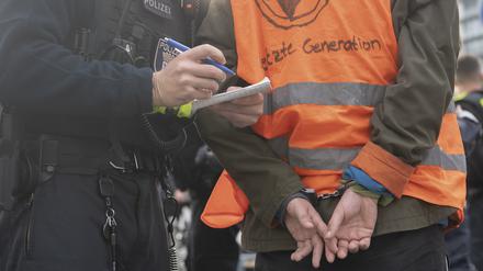 Ein Aktivist der Klimaschutz-Gruppe Letzte Generation steht am Potsdamer Platz in Handschellen neben einem Polizeibeamten.