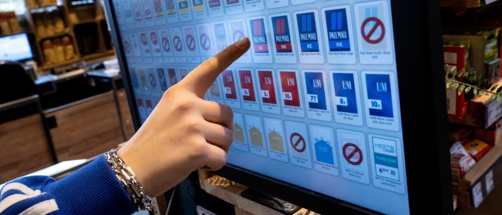 Eine Frau tippt an der Kasse eines Supermarktes mit dem Finger auf das Display von einem Zigarettenautomaten.