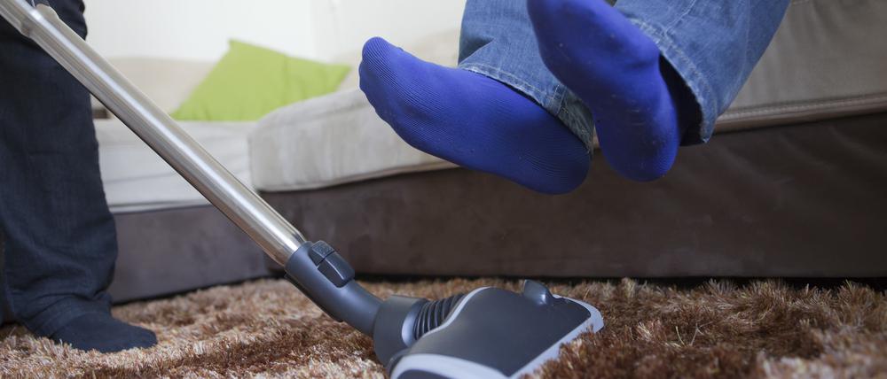 Ein Mann hebt seine Beine, während eine Frau den Teppich saugt.