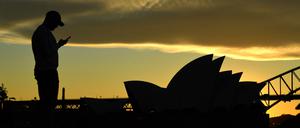 Ein Mann steht bei Sonnenuntergang vor dem Sydney Opera House und blickt auf sein Mobiltelefon. (Archivbild).