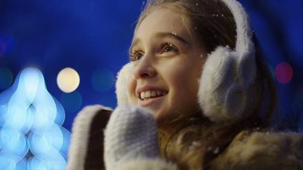 Close up of the happy cute girl looking charmed at something and clapping in white winter gloves on the blurred Christmas lights in the night. Portrait shot. Outside