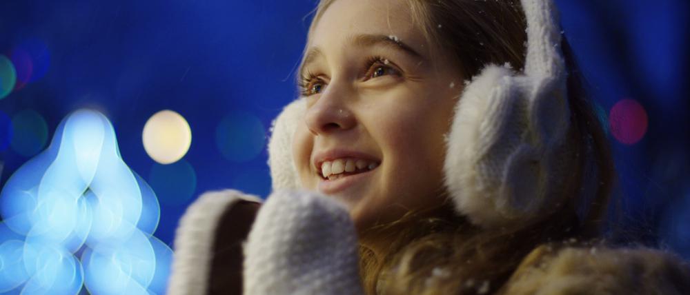 Close up of the happy cute girl looking charmed at something and clapping in white winter gloves on the blurred Christmas lights in the night. Portrait shot. Outside