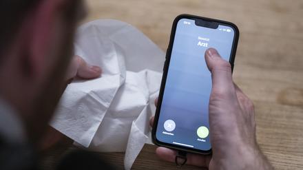Ein Mann hält ein Taschentuch in einer Hand und ein Telefon mit dem Schriftzug „Arzt“ in der anderen. 