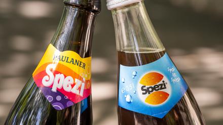 Eine Flasche des Kultgetränks „Spezi“ (l) von der Großbrauerei Paulaner und eine Flasche „Spezi“ der Augsburger Riegele Brauerei werden nebeneinander gehalten. 