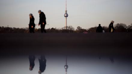 Spaziergänger gehen auf dem Tempelhofer Feld vor dem Berliner Fernsehturm, wobei sich die Szenerie in einer Pfütze spiegelt.