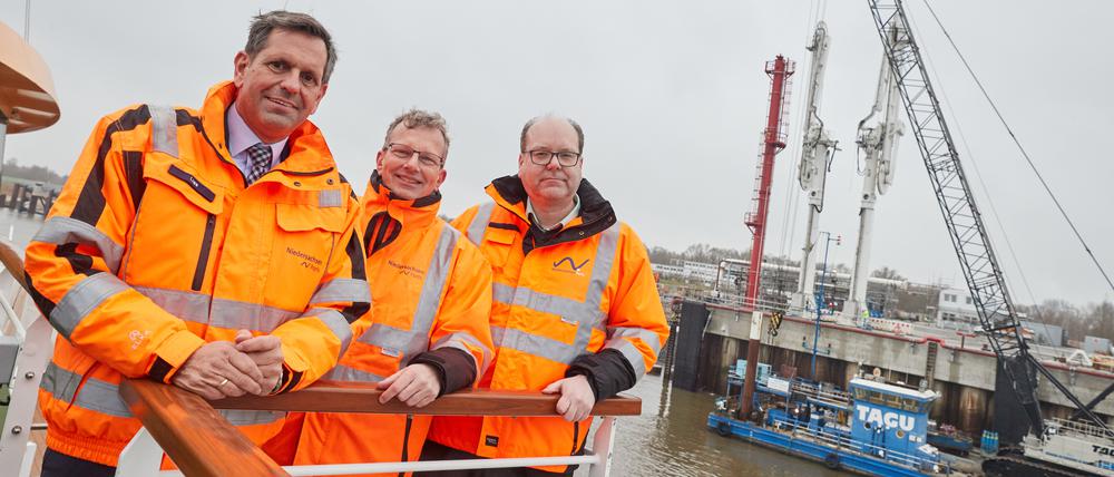 Olaf Lies (SPD), Holger Banik und Christian Meyer (Die Grünen) stehen auf der MS Helgoland. Im Hintergrund ist der Anleger für verflüssigte Gase (AVG) mit seinen Gas-Löscharmen zu sehen.