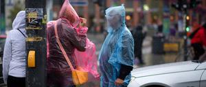 Passanten mit Regenschutzhüllen stehen in Berlin auf der Straße am Potsdamer Platz.. Es wird regnerisch und windig.