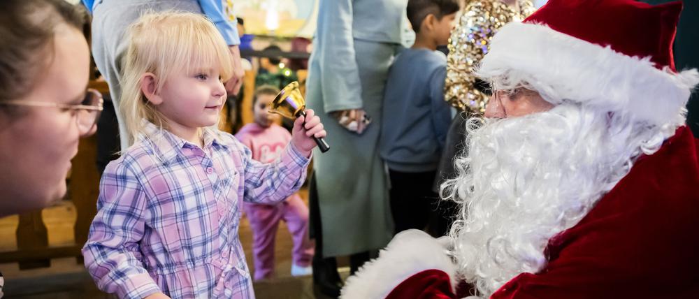 24.12.2023, Berlin: Die zweijährige Amelia-Marie läutet bei einem kostenlosen Weihnachtessen der Arche und des Hofbräus Berlin für Kinder und Familien aus benachteiligten Verhältnissen im Hofbräu Wirtshaus mit der Glocke des Weihnachtsmannes, nachdem sie von ihm ein Geschenk bekommen hat. Foto: Christoph Soeder/dpa - ACHTUNG: Nur zur redaktionellen Verwendung im Zusammenhang mit der aktuellen Berichterstattung über das genannte Thema und nur mit vollständiger Nennung des vorstehenden Credits +++ dpa-Bildfunk +++