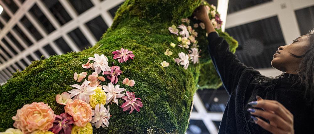 Eine Frau befestigt in der Blumenhalle auf der Internationalen Grünen Woche verschiedene Blumen an der Moos-Skulptur eines Bären.  