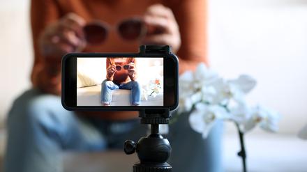 Eine Frau sitzt mit einer Sonnenbrille in Händen auf einem Sofa und filmt sich dabei mit ihrem iPhone (gestellte Szene). Produkttipps auf Instagram oder Tiktok sind oft bezahlte Werbung.