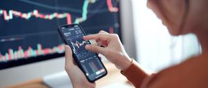 Frau blickt auf ihr Smartphone, auf dem Aktiencharts abgebildet sind.