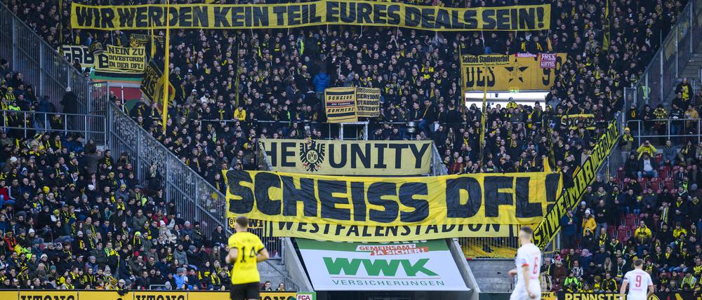 Fußball: Bundesliga, FC Augsburg - Borussia Dortmund, 15. Spieltag, WWK-Arena. Fans von Borussia Dortmund halten ein Plakat mit der Aufschrift „Wir werden kein Teil eures Deals sein! Scheiss DFL“ in die Luft. Das Fan-Bündnis «Unsere Kurve» fordert eine Wiederholung der Abstimmung beim Investoren-Deal. (zu dpa: «Investoren-Deal: Fan-Bündnis fordert Wiederholung der Abstimmung») +++ dpa-Bildfunk +++