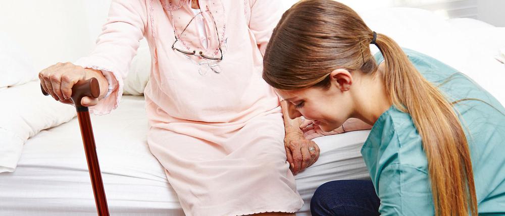 Altenpfleger hilft Seniorin beim Aufstehen und Anziehen der Hausschuhe im Pflegeheim.