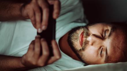 Ein junger Mann liegt im Bett und schaut auf ein Smartphone.
