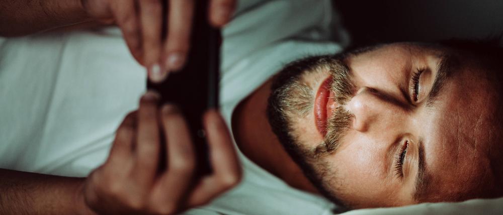 Ein junger Mann liegt im Bett und schaut auf ein Smartphone.