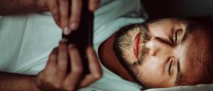 Ein junger Mann liegt im Bett und schaut auf ein Smartphone.