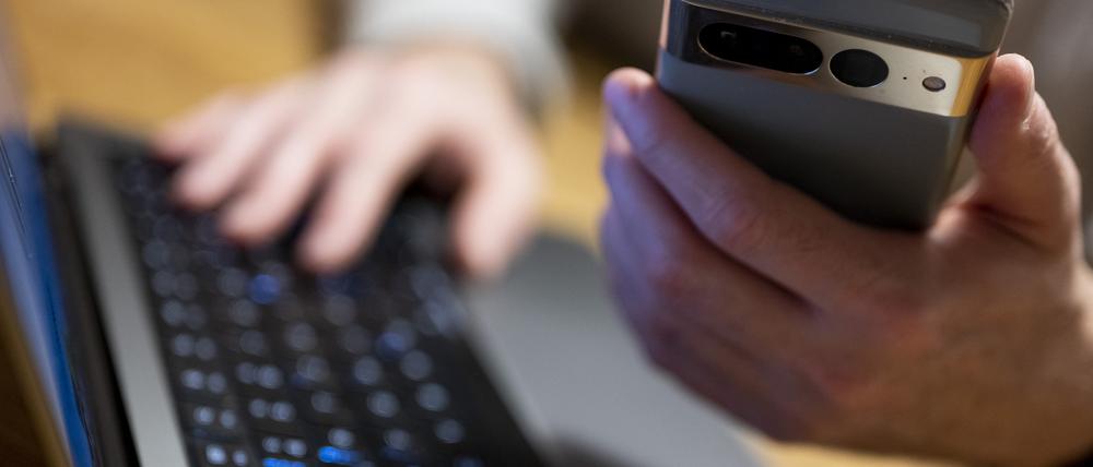 Ein Mann sitzt am Laptop und hält ein Smartphone mit in der Hand. (Symbolbild)