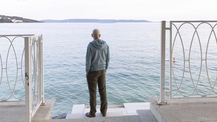 Mann steht am Meer unter einer Pinie und guckt in die Ferne.