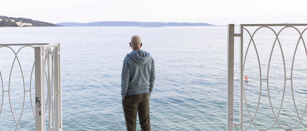 Mann steht am Meer unter einer Pinie und guckt in die Ferne.