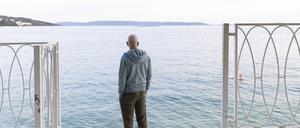 Mann steht am Meer unter einer Pinie und guckt in die Ferne.