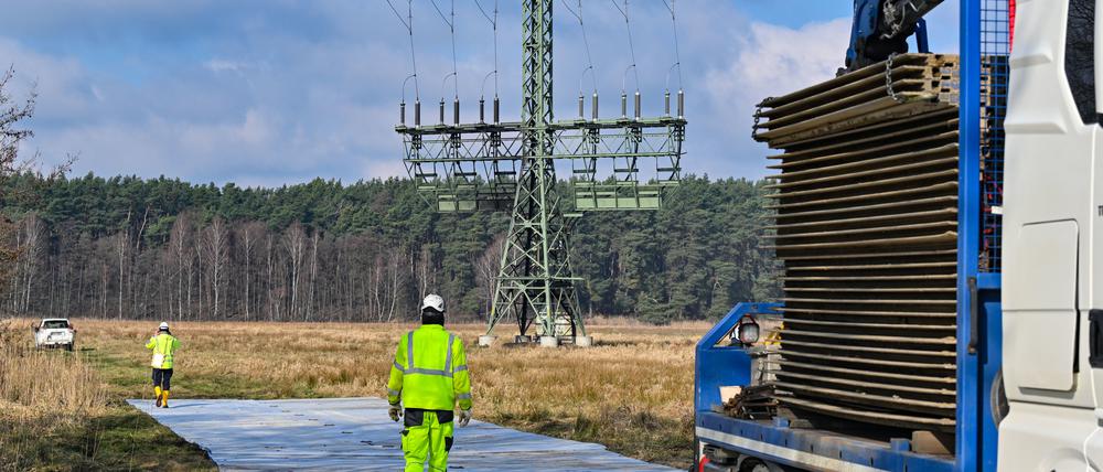 06.03.2024, Brandenburg, Grünheide: Arbeiter errichten eine provisorische Baustraße zu einem Strommast mit Brandspuren auf einem Feld nahe der Tesla-Autofabrik.  