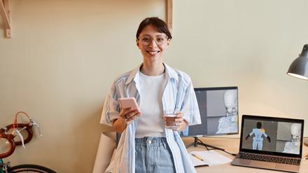 Eine lächelnde junge Frau mit Smartphone und Laptop am digitalen Arbeitsplatz.