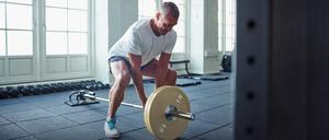 Ein älterer Mann trainiert mit einer Langhantel in einem Fitnessstudio seine Rückenmuskulatur.