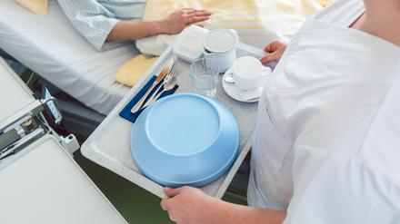 Eine Krankenpflegerin bringt ein Tablett mit Essen ans Bett einer Patientin.
