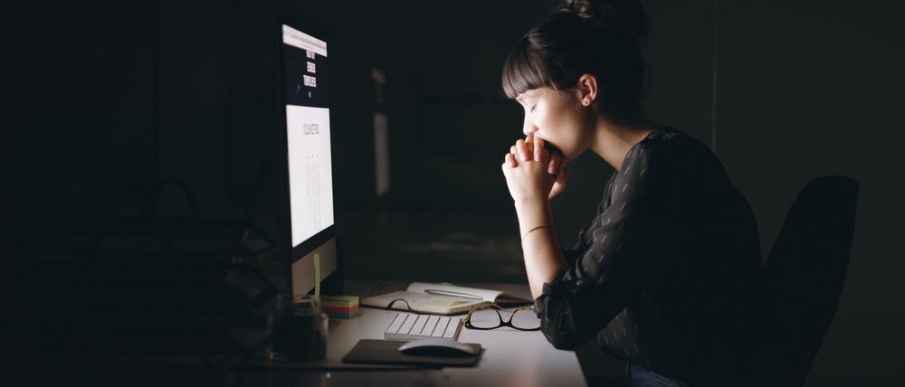 Shot of a young businesswoman sitting with her hands clasped while working on a computer in an office at night