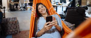 Happy businesswoman holding smart phone lying in hammock in office