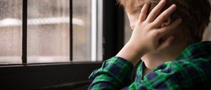 Ein kleiner Junge sitzt traurig vor dem Fenster und hält sich die Ohren zu.