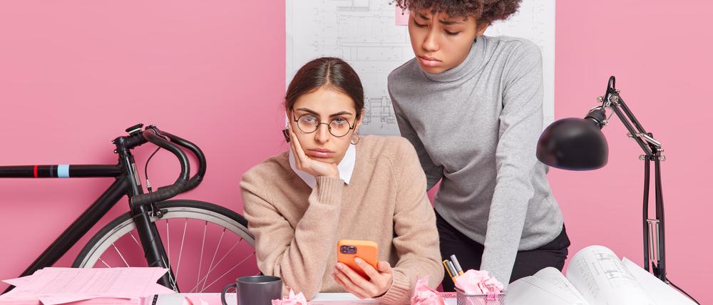 Zwei junge Arbeitnehmerinnen schauen gelangweilt auf ein Handy.