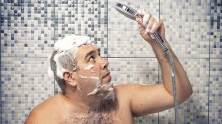 Ein Mann steht mit Shampoo im Haar unter der Dusche und blickt unsicher auf den Duschkopf in seiner Hand.