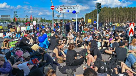 11.05.2024, Brandenburg, Grünheide: Viele Teilnehmer einer Protestveranstaltung sitzen auf einer sitzen und stehen auf einer Straße vor dem Werk von Tesla. Nach teils heftigen Auseinandersetzungen am 10.05.2024 am Tesla-Werksgelände in Grünheide bei Berlin stellt laufen derzeit neue Proteste gegen das US-Unternehmen. Foto: Patrick Pleul/dpa +++ dpa-Bildfunk +++