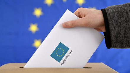 Ein Wahlbrief wird in eine Wahlurne gesteckt, im Hintergrund ist eine EU-Flagge zu sehen.