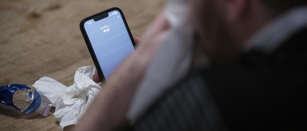 Ein Mann hält ein Taschentuch in einer Hand und ein Telefon mit dem Schriftzug „Arzt“ in der Anderen.