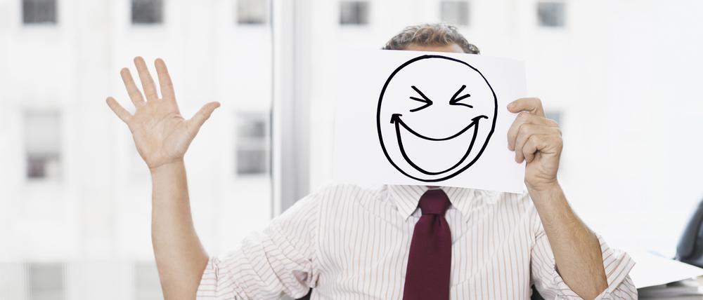 Ein Geschäftsmann hält ein Papier mit einem gemalten, breit grinsenden Smiley vor seinem Gesicht.