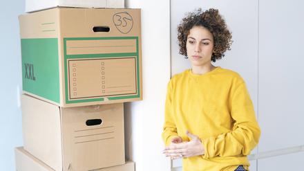Eine nachdenklich wirkende Frau steht in einer Wohnung neben gepackten Kisten. 