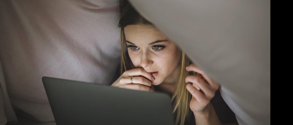 Eine junge Frau schaut unter der Bettdecke liegend auf den Bildschirm ihres Laptops.