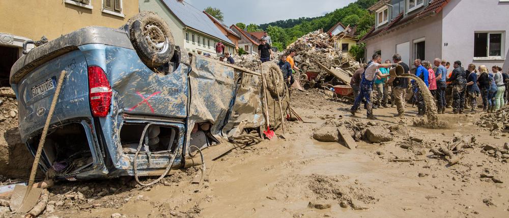 Helfer bilden eine Menschenkette, um möglichst viel Schlamm mit Eimern aus einem Haus im Ort zu bekommen, während davor ein Auto auf dem Dach liegt.
