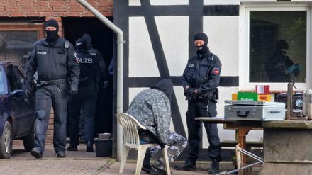 Polizisten sind bei einer Razzia im Einsatz, während ein Mann in Kleidung mit Tarnfleckmuster auf einem Stuhl vor einem Polizisten sitzt. 