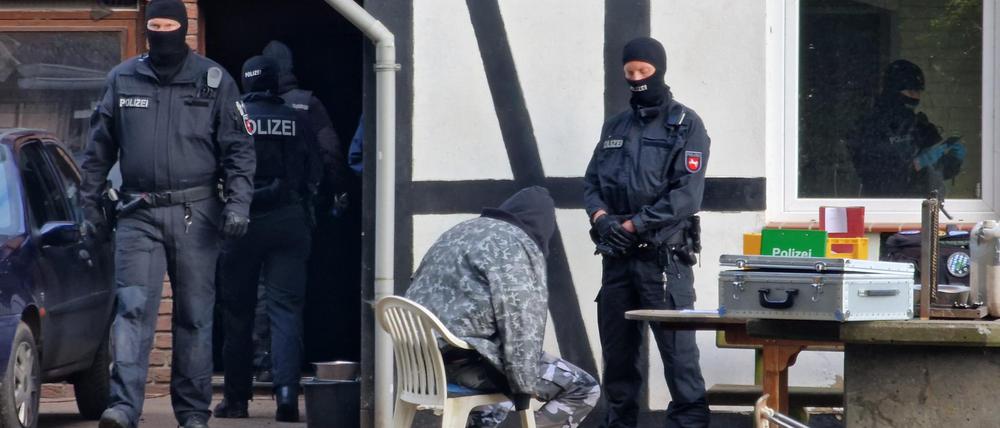 Polizisten sind bei einer Razzia im Einsatz, während ein Mann in Kleidung mit Tarnfleckmuster auf einem Stuhl vor einem Polizisten sitzt. 